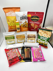 A variety of packaged snacks and protein products are arranged on a white surface, including cheese balls, crunch mixes, pizza crisps, oatmeal, and flavored drinks, showcasing healthy and indulgent options.