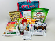 A colorful assortment of protein snacks, including chips, macaroons, and pancakes, is displayed in front of a blue bag with "syntax" written in pink, set against a plain white background.