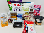 A variety of protein-rich snacks and drinks, including kale chips, beef jerky, protein powders, and a hot chocolate mix, are displayed on a white background, showcasing their packaging and nutritional information.