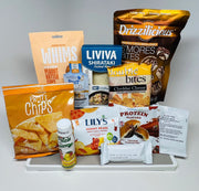 A variety of snack products is displayed on a white surface, including peanut butter cups, shirataki rice, cheddar cheese bites, gummy bears, and protein drinks, all arranged for an appealing presentation.