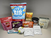 A variety of snack products is displayed on a neutral background, including puffcorn, protein crisps, oatmeal, muffins, and protein bars, showcasing their packaging and nutritional highlights.