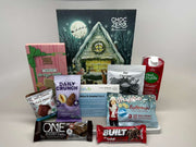 A collection of various snacks and beverages, including keto chocolate, protein bars, and hot chocolate, arranged in front of a festive winter-themed backdrop featuring a cozy house and Santa.