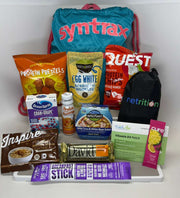 A colorful assortment of health and protein snacks, including protein chips, oatmeal, and a vitamin patch, is displayed in front of a bright blue Syntrax drawstring bag on a white background.