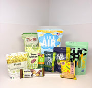 A variety of healthy snacks and food products, including popcorn, chips, and bars, are arranged on a white background, showcasing brands like Bob's Red Mill and Poshi.
