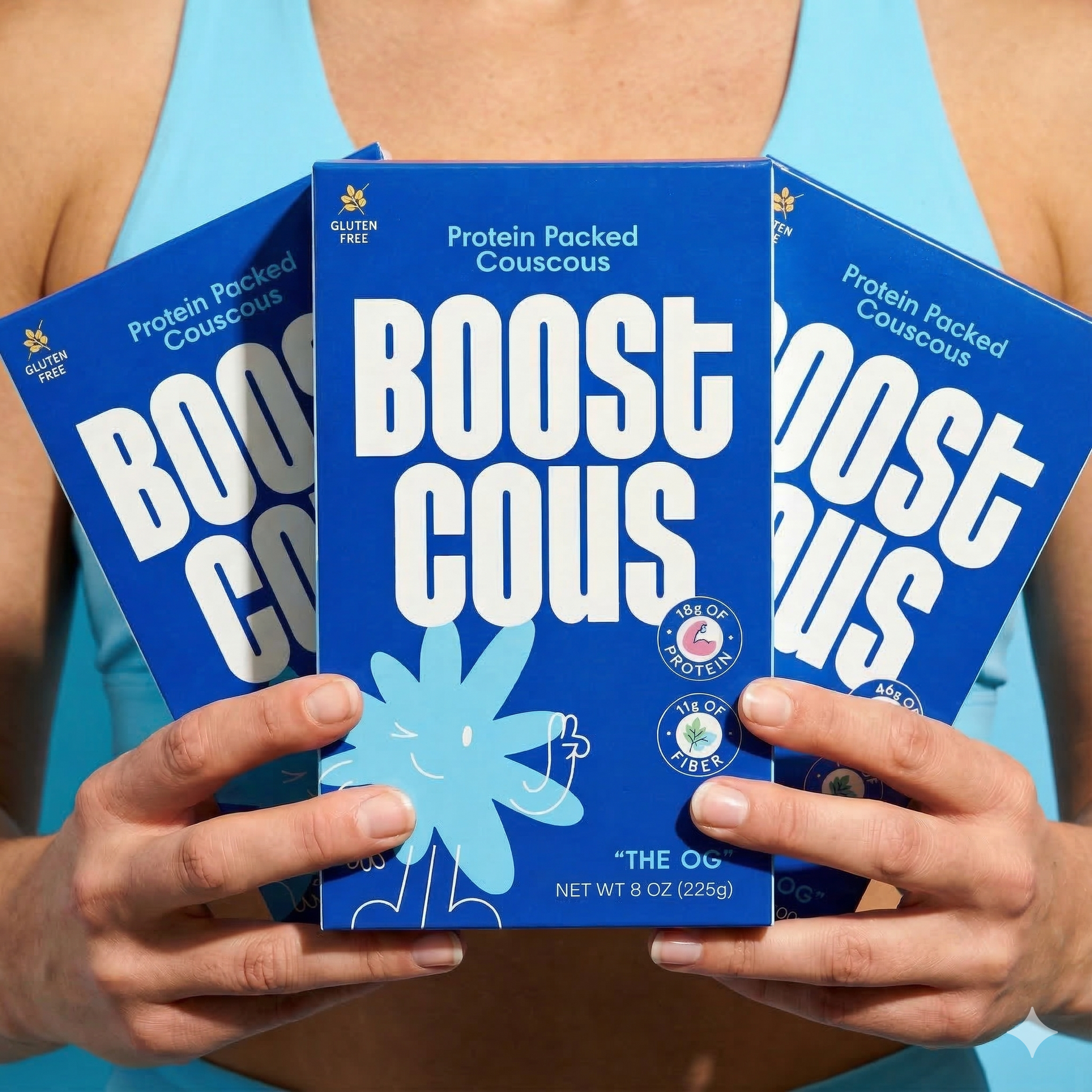 A person holds three boxes of "Boost Couscous," a protein-packed, gluten-free product, against a light blue background, showcasing its vibrant packaging and nutritional highlights.