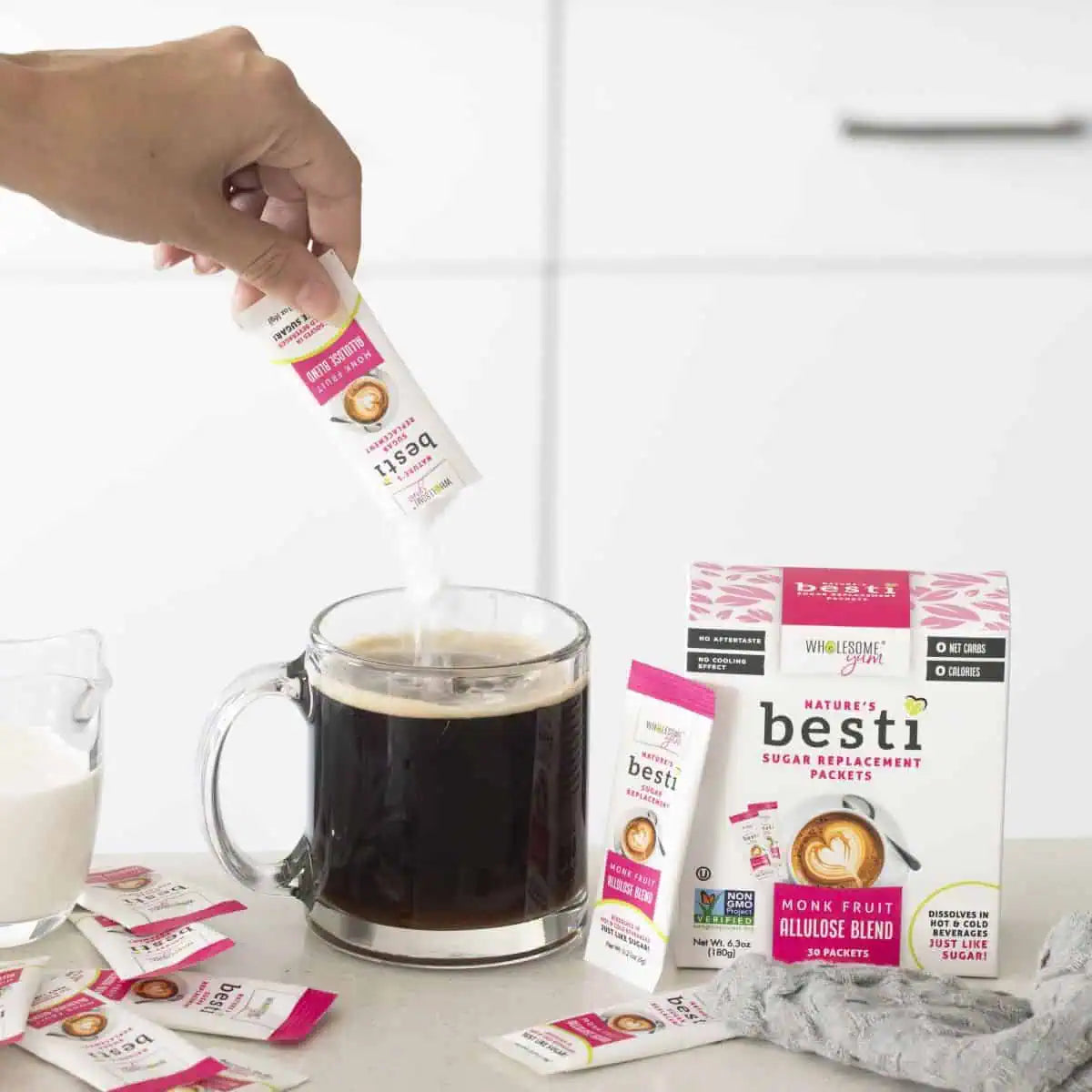 A hand is pouring a sugar replacement packet into a glass of black coffee, with a box of Nature's Besti sugar replacement and a jug of cream nearby on a light countertop.