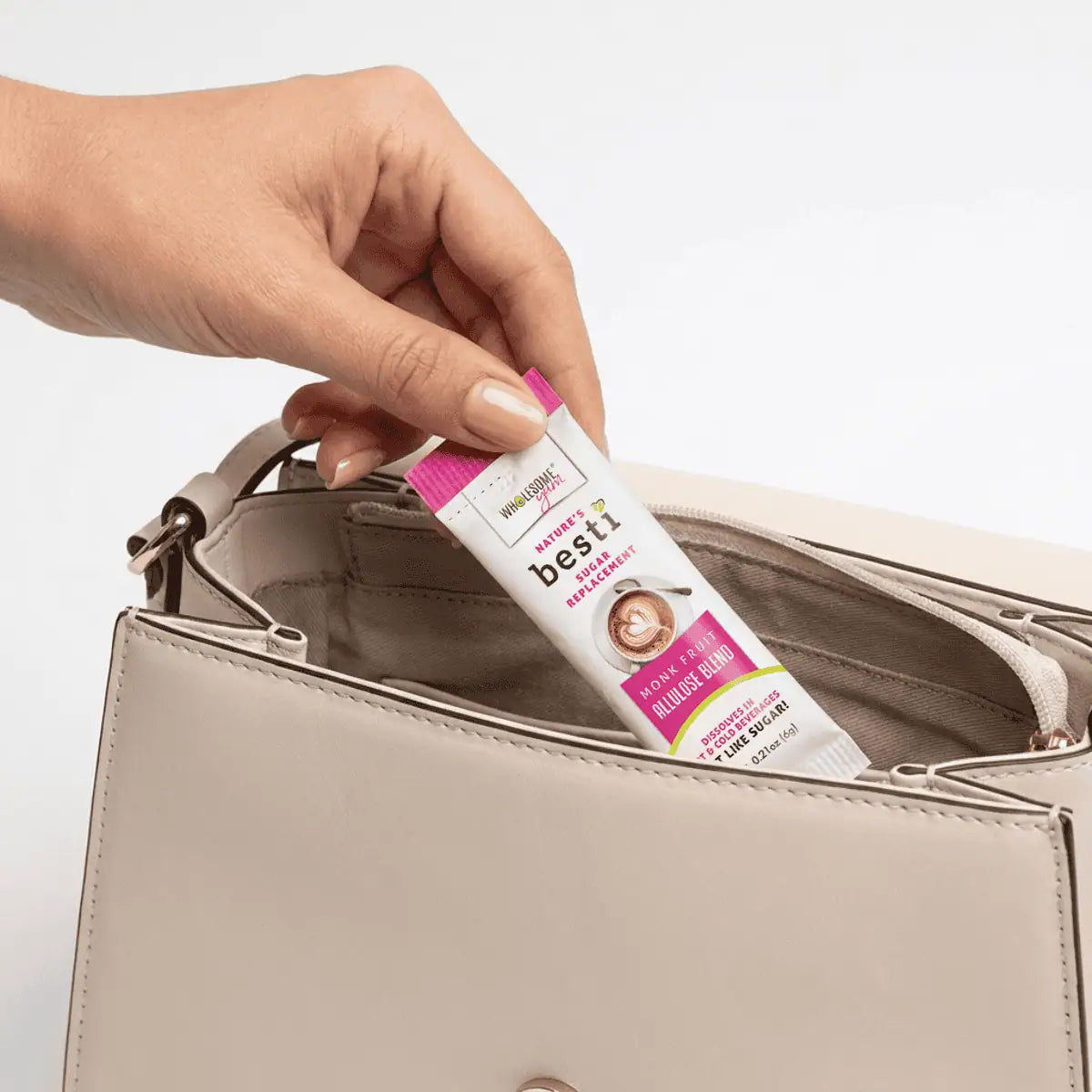 A hand is placing a packet of Nature's Besti sugar replacement into the pocket of a light-colored handbag against a plain background.