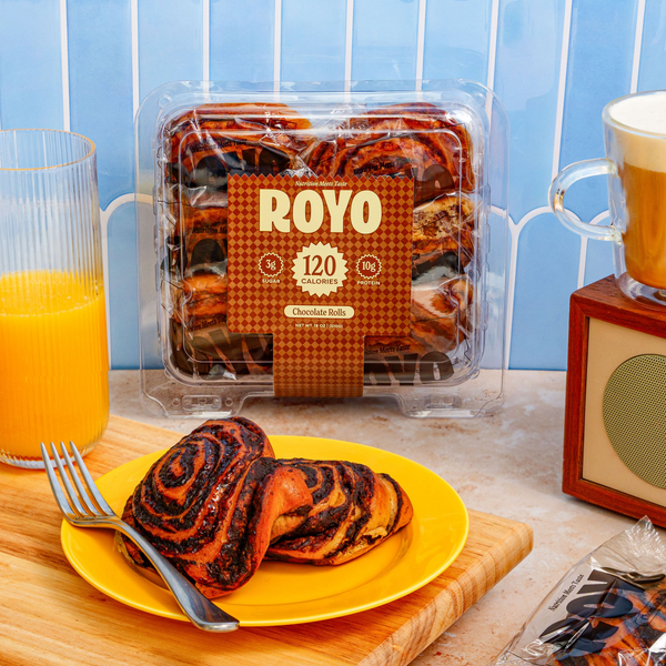 A yellow plate holds two chocolate rolls, with a fork beside it, while a clear package of Royo chocolate rolls and a glass of orange juice sit on a wooden cutting board against a light blue background.