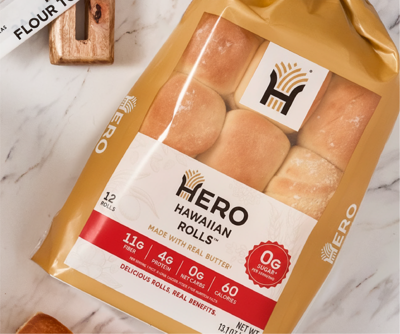 A package of Hero Hawaiian Rolls, featuring twelve soft rolls made with real butter, is displayed on a marble surface, highlighting nutritional information and branding.