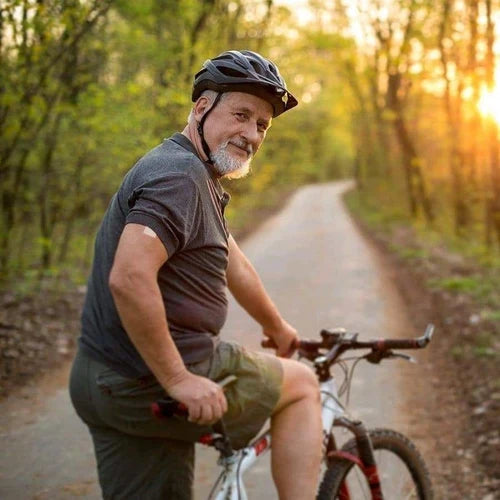 An older man with a gray beard, wearing a helmet and casual clothing, smiles while sitting on his mountain bike on a tree-lined path during sunset.