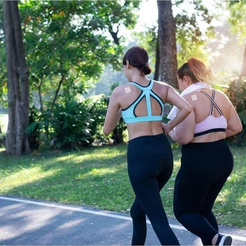 Two women jog together on a sunny path in a park, wearing athletic tops and leggings, with visible sweat patches and supportive posture, enjoying their workout in a natural setting.